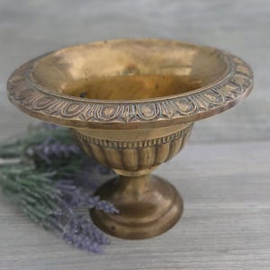May include: A brass pedestal bowl featuring a wide, flared rim with a decorative scalloped edge pattern. The bowl has a fluted, ribbed body that tapers down to a circular base. The metal surface shows a warm, aged patina with subtle dark highlights.