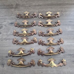 May include: A collection of ten brass drawer pulls arranged in five rows of two. Each pull features a curved handle attached to a decorative backplate with scalloped edges and a central finial. The metal has a warm, aged patina finish.