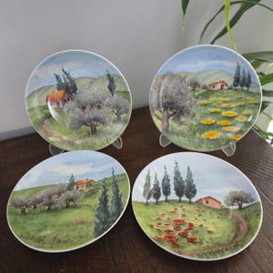 Puede incluir: Cuatro platos decorativos con pinturas de paisajes. Cada plato presenta una escena de acuarela diferente de un paisaje rural con árboles, casas y caminos sinuosos. La paleta de colores incluye verdes, azules y tonos tierra.