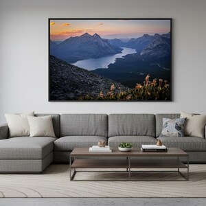 Pode incluir: Grande impressão artística emoldurada com uma paisagem de montanha, um lago e um pôr do sol. A obra de arte está pendurada acima de um sofá seccional cinza com almofadas decorativas e uma mesa de centro de madeira. O quarto tem uma paleta de cores neutras.