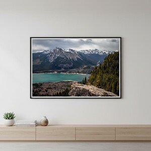 Pode incluir: Fotografia emoldurada de uma paisagem de uma cordilheira com picos cobertos de neve, um lago turquesa e uma floresta. A fotografia está em uma moldura preta e é exibida em um armário de madeira clara.