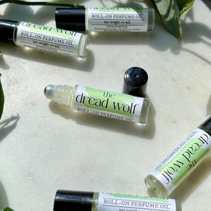 May include: Several clear glass roll-on perfume oil bottles with black caps are scattered on a white surface. The labels read "the dread wolf" and "roll-on perfume oil." The labels also include the net weight of 10 mL and ingredients.