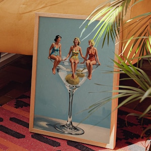 May include: A framed print featuring three women in vintage swimsuits perched on the rim of a giant martini glass with an olive. The artwork has a light blue background and is set against a warm, inviting interior with a patterned rug and a yellow chair.