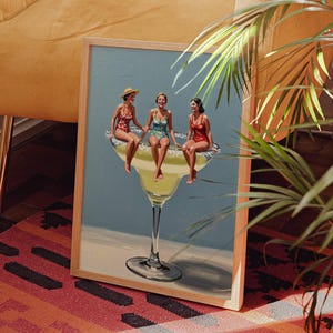 May include: A framed art print depicts three women in vintage swimsuits seated on a giant margarita glass. The artwork has a light blue background and is set against a warm, inviting interior with a yellow chair and a patterned rug.