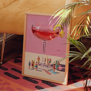 May include: A framed art print featuring a giant cocktail glass filled with a red liquid and a lemon twist. Miniature figures of people with surfboards and beach attire are gathered around the glass, set against a pink background.