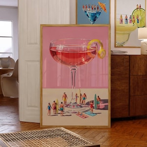 May include: A framed wall art print featuring a large pink cocktail in a stemmed glass with a lemon twist garnish. Below the glass, miniature figures in swimwear stand on a sandy surface with surfboards and towels, creating a surreal beach scene.