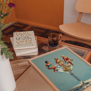 May include: A framed print shows three women relaxing in a giant martini glass with olives. The artwork is on a glass-topped table with books and a coffee mug. A white vase with purple flowers is in the background.