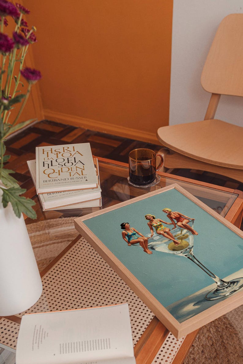 May include: A framed print shows three women relaxing in a giant martini glass with olives. The artwork is on a glass-topped table with books and a coffee mug. A white vase with purple flowers is in the background.