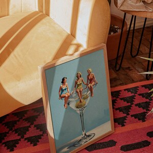 May include: A framed print depicting a vintage illustration of three women in swimsuits sitting on a large martini glass with olives. The artwork has a light blue backdrop and is set in a wooden frame.