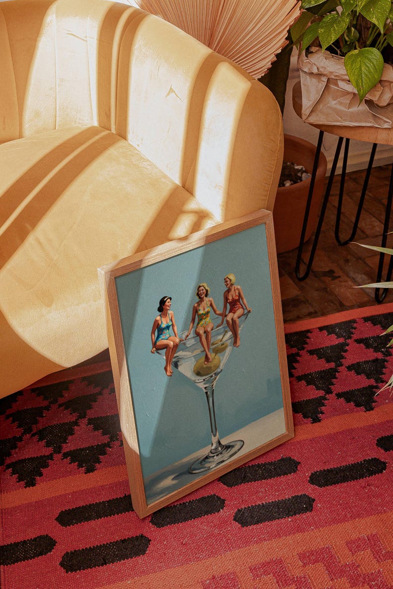 May include: A framed print depicting a vintage illustration of three women in swimsuits sitting on a large martini glass with olives. The artwork has a light blue backdrop and is set in a wooden frame.