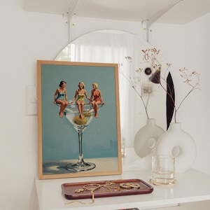 May include: A framed print shows three women in vintage swimsuits perched on a large martini glass with an olive. The artwork is on a white shelf with a mirror, vases, and a tray of jewellery. Other items include a woven basket.