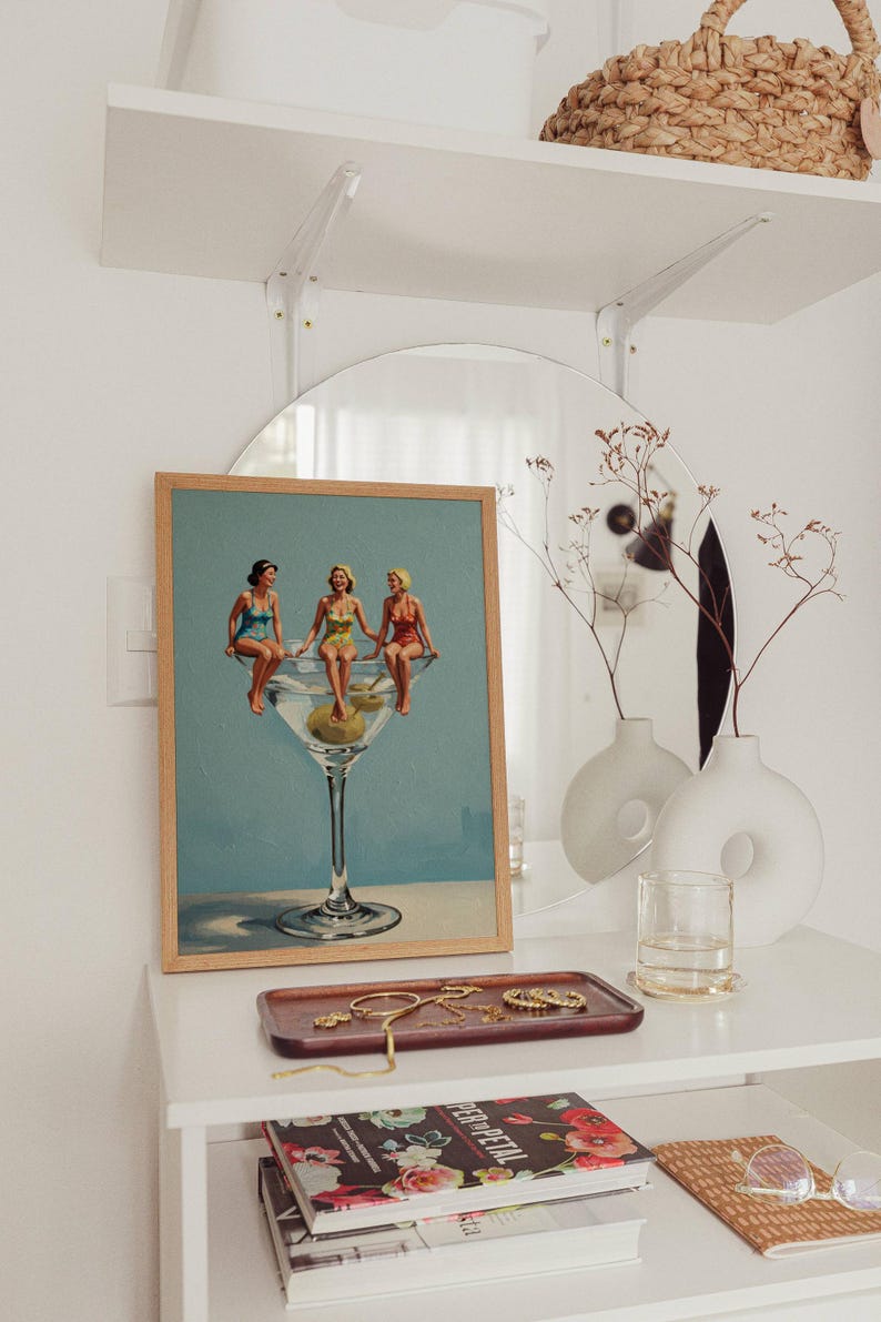 May include: A framed print shows three women in vintage swimsuits perched on a large martini glass with an olive. The artwork is on a white shelf with a mirror, vases, and a tray of jewellery. Other items include a woven basket.