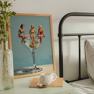 May include: A framed print shows three women in swimsuits sitting on a giant martini glass with olives. A clear glass vase with flowers and a textured white mug on a wooden coaster sit on a mint green bedside table next to a black metal bed frame.