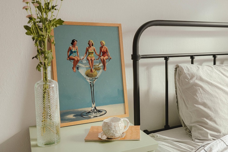 May include: A framed print shows three women in swimsuits sitting on a giant martini glass with olives. A clear glass vase with flowers and a textured white mug on a wooden coaster sit on a mint green bedside table next to a black metal bed frame.