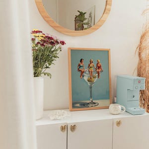 May include: A framed print shows three women in a martini glass. A light blue espresso machine and a white mug sit on a white cabinet. A round wooden mirror hangs above the cabinet, and a vase of flowers is to the left.