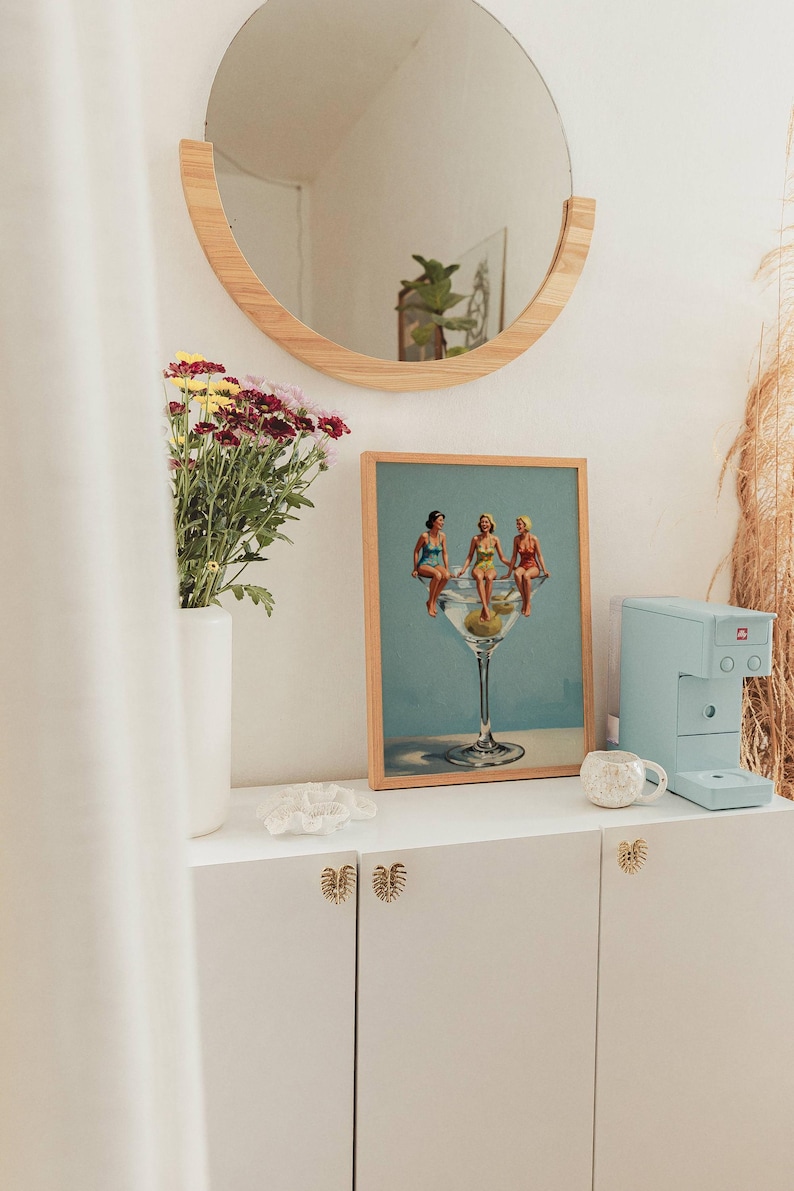 May include: A framed print shows three women in a martini glass. A light blue espresso machine and a white mug sit on a white cabinet. A round wooden mirror hangs above the cabinet, and a vase of flowers is to the left.