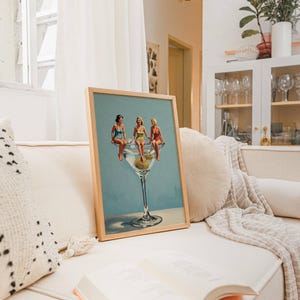May include: A framed print shows three women in swimsuits perched on a giant martini glass with olives. The artwork has a light blue background and is displayed on a cream sofa with a book. The frame is made of wood.