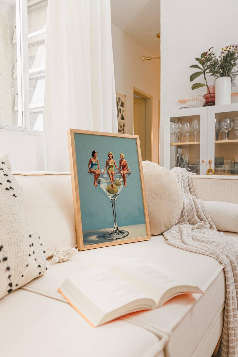 May include: A framed print shows three women in swimsuits perched on a giant martini glass with olives. The artwork has a light blue background and is displayed on a cream sofa with a book. The frame is made of wood.