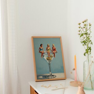 May include: A framed print showing three women in vintage swimsuits perched on a giant martini glass with olives. The artwork has a light blue background and is displayed on a white surface with a vase and candle.