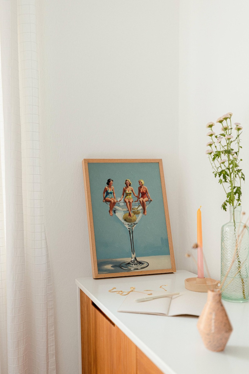 May include: A framed print showing three women in vintage swimsuits perched on a giant martini glass with olives. The artwork has a light blue background and is displayed on a white surface with a vase and candle.