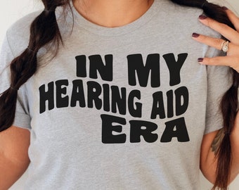 May include: A gray t-shirt with the text "In My Hearing Aid Era" printed in black. The text is in a retro, bold font.