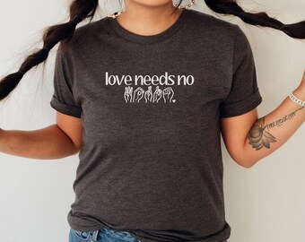 May include: A dark gray t-shirt with the text "love needs no" and a graphic of the word "sorry" in American Sign Language.