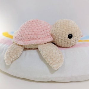 May include: A crocheted plush toy turtle with a pink shell and beige body. The turtle is lying on its side on a white and pink patterned surface.