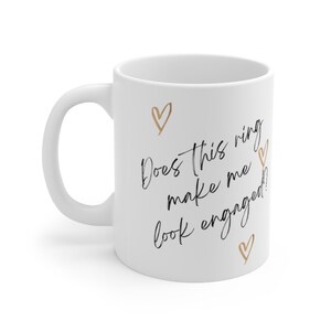 May include: White ceramic mug with the text "Does this ring make me look engaged?" in black script. There are four gold hearts around the text.