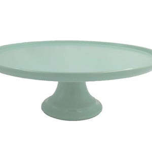 May include: A solid light mint green cake stand featuring a wide, flat circular top surface with a slightly raised rim and a sturdy, flared pedestal base. The entire structure is finished in a smooth, uniform matte color throughout the piece.