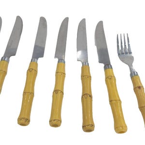 May include: A set of eight pieces of flatware featuring silver-toned metal heads and light brown, textured bamboo-style handles. The collection includes six knives with serrated edges, one four-tined fork, and one spoon, all arranged in a row.