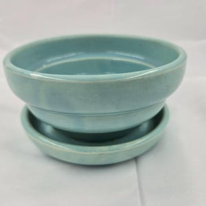 May include: A light blue ceramic bowl with a matching saucer. The bowl features a smooth, glossy finish with subtle tonal variations in the glaze, a rounded rim, and a slightly tapered base that rests securely within the circular, shallow saucer.