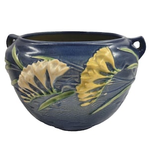 May include: A deep blue ceramic bowl featuring raised relief designs of cream-colored flowers and green leaves. The bowl has a rounded body, a wide opening, and two small handles on opposite sides. The surface texture includes wavy lines.