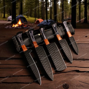 May include: A set of six black chef knives with wooden handles and textured blades, displayed on a wooden surface. The knives are arranged next to a black leather knife roll. The background features a forest scene with tents and a campfire.