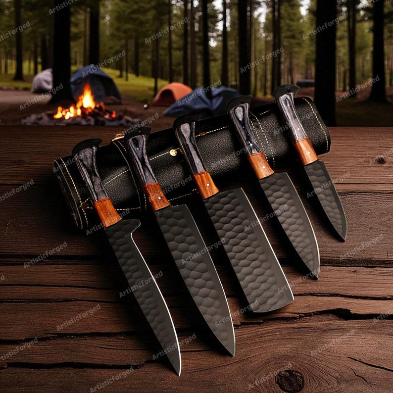 May include: A set of six black chef knives with wooden handles and textured blades, displayed on a wooden surface. The knives are arranged next to a black leather knife roll. The background features a forest scene with tents and a campfire.