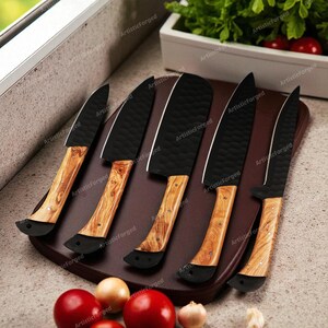 May include: A set of six kitchen knives with black blades and wooden handles, arranged on a brown cutting board. The handles have a light wood grain pattern. Tomatoes, onions, and garlic are in the foreground. The text "ArtisticForged" is visible.
