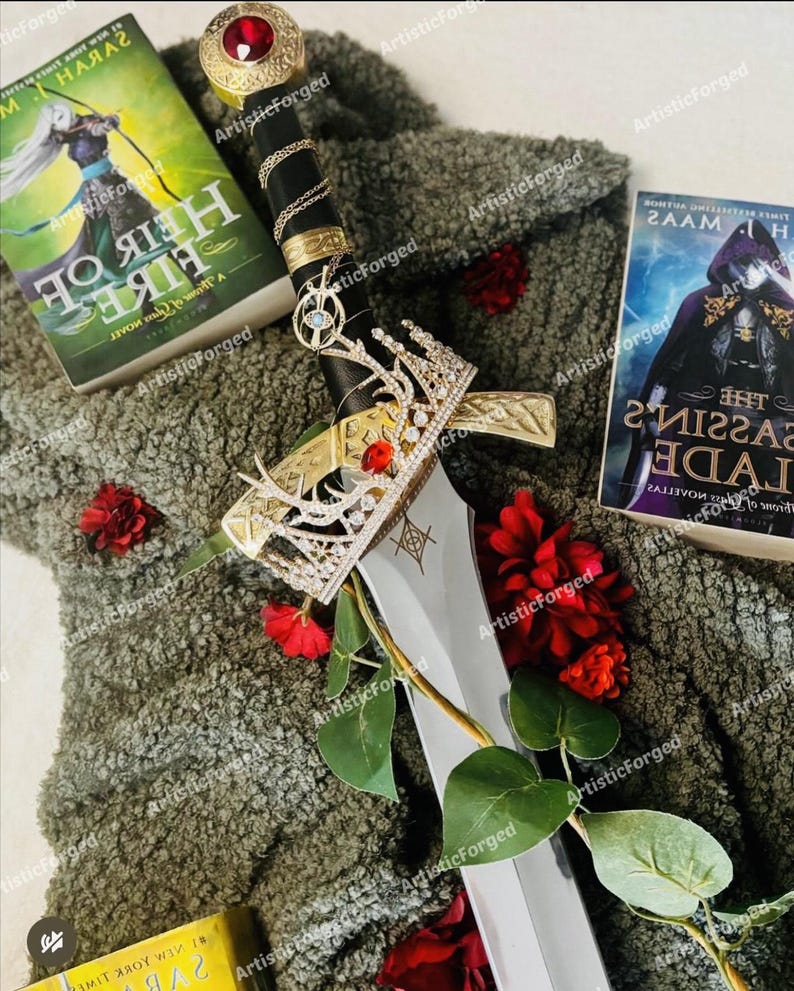 May include: A silver sword with a gold hilt and a red jewel on top. The sword is lying on a green fuzzy surface with red flowers and two books. The books are titled "A Heir of Fire" and "The Assassin's Blade".