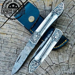 May include: Two ornate pocket knives with intricate floral designs on the blades. The blades are made of Damascus steel and have a polished finish. The knives are in a leather sheath.