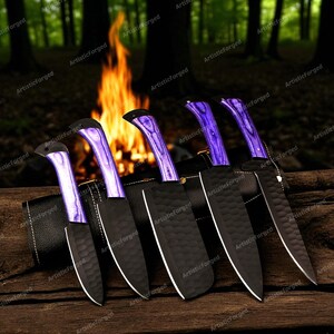May include: A set of six black knives with purple handles and black sheaths. The blades have a textured finish and white edges. The knives are displayed on a wooden surface with a blurred background of a campfire and trees.
