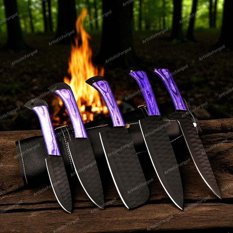 May include: A set of six black knives with purple handles and black sheaths. The blades have a textured finish and white edges. The knives are displayed on a wooden surface with a blurred background of a campfire and trees.