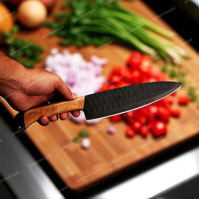 May include: A black chef's knife with a textured blade and a light wood handle. The knife is held over a wooden cutting board with chopped vegetables, including tomatoes and onions. The blade has a hammered finish.