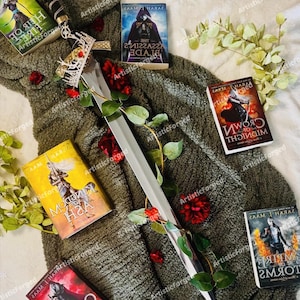 May include: A silver sword with a gold crown handle, lying on a green blanket with red roses. There are several fantasy novels with titles in a foreign language, including "The Assassin's Blade", "Crown of Midnight", "Queen of Shadows", and "Empire of Storms".