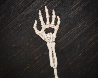 May include: A bottle opener featuring a handle shaped like a human forearm and a head shaped like a skeletal hand. The bone-colored structure has dark, weathered detailing in the crevices, with the palm area forming the functional opening mechanism.