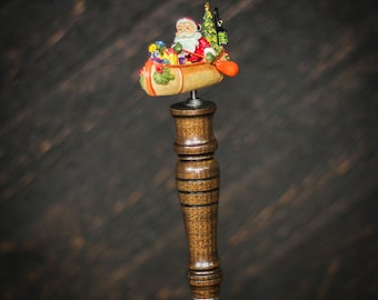 May include: A wooden bottle stopper with a Santa Claus figurine in a canoe. The canoe is filled with presents and a Christmas tree. The stopper is brown with a dark wood grain.