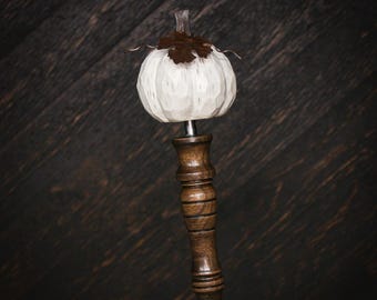 May include: A bottle stopper featuring a carved white pumpkin topper with a dark brown leaf accent. The pumpkin is mounted on a turned dark brown wooden handle with horizontal grooves, which connects to a silver-toned metal base component.
