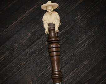 May include: A dark brown wooden tap handle featuring a carved off-white figure of a skeleton wearing a wide-brimmed hat and a poncho. The figure sits atop a cylindrical, tiered wooden base that transitions into a smooth metal ferrule at the bottom.
