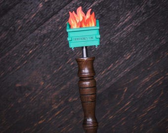 May include: A beer tap handle featuring a dark brown wooden grip and a teal dumpster topper with orange and red flames emerging from the top. The dumpster has the text EVERYTHING'S FINE printed in black on the front side of the container.