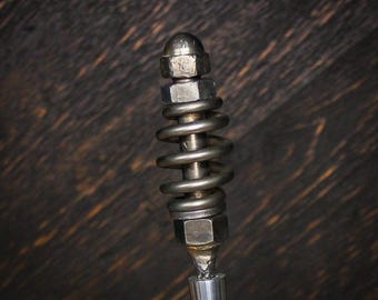 May include: A metallic handle featuring a central coiled spring mechanism secured between two hexagonal nuts and a rounded top cap. The assembly is mounted onto a smooth, tapered silver-colored shaft, showcasing a raw industrial aesthetic.
