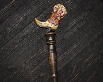 May include: A decorative tap handle featuring a miniature cowboy boot topper. The boot is painted in cream and red with gold floral detailing. The handle base is made of dark brown wood with a cylindrical shape and a silver metal ferrule at the bottom.