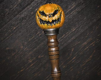 May include: A bottle stopper featuring a carved orange pumpkin head with a menacing black facial expression. The pumpkin is mounted on a dark brown, turned wooden handle with horizontal grooves, connected by a silver-toned metal collar.