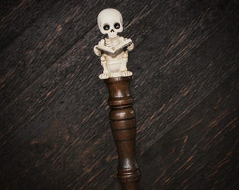 May include: A decorative handle featuring a cream-colored skeleton figurine sitting on a toilet while reading an open book. The figurine is mounted atop a dark brown, turned wooden handle with a smooth, polished finish and a metallic base.
